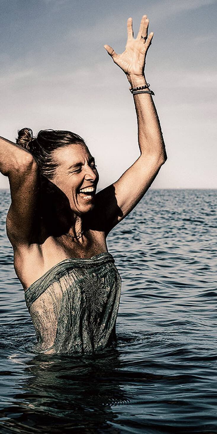 living inquiry by tomm claude Eine lächelnde Frau steht hüfttief im Meer und hält spielerisch ein Kleidungsstück hoch, während sie in die Kamera schaut. Sie verkörpert innere Ruhe und Freude daran, im Hier und Jetzt zu sein, während sich hinter ihr der Himmel und der Horizont erstrecken. spirituelles coaching und zen meditation für emotionale heilung, erwachen und essenz