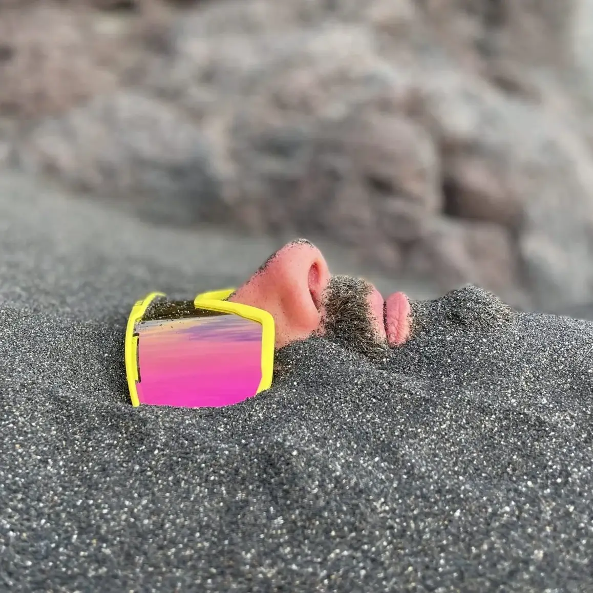 living inquiry by tomm claude Ein Mann liegt im Sand eingegraben, nur seine Nase, sein Mund, sein Bart und seine reflektierende gelbe Sonnenbrille sind über der Oberfläche sichtbar - eine ruhige Szene, die an spirituelles Coaching und Zen-Meditation für emotionale Heilung und Verkörperung erinnert. Sandige Felsen bilden den Hintergrund. spirituelles coaching und zen meditation für emotionale heilung und embodiment