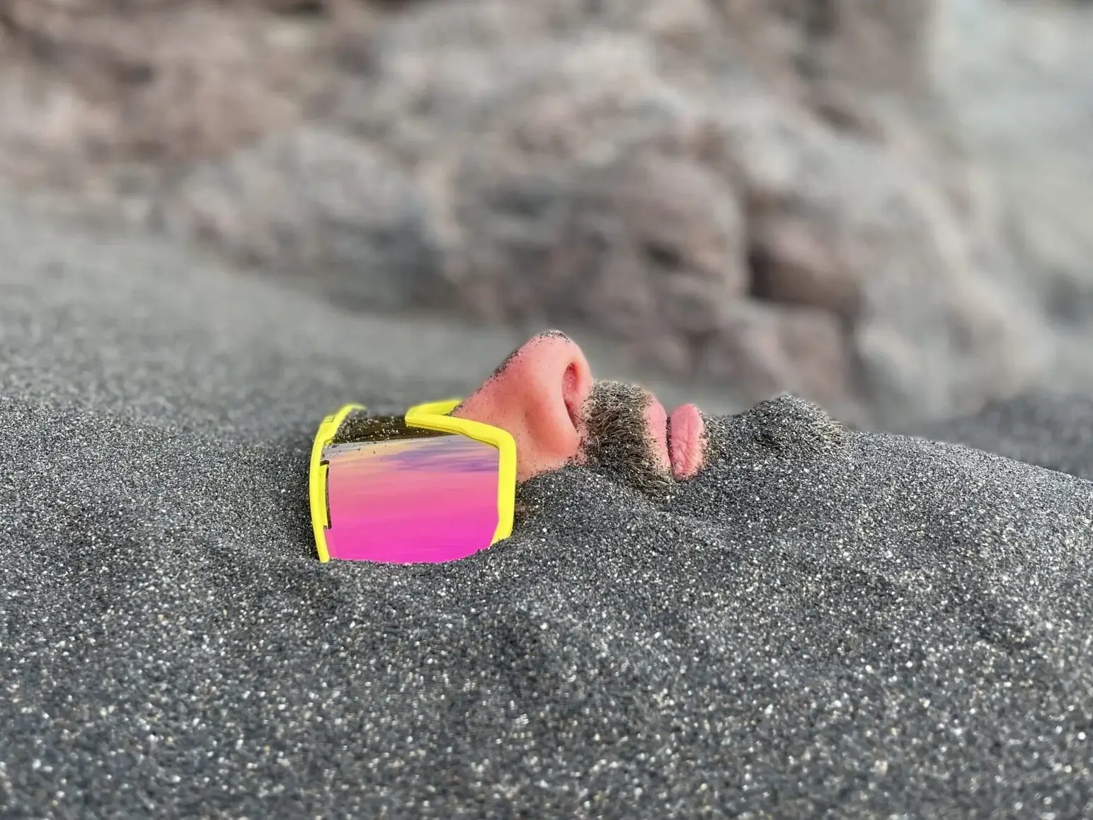living inquiry by tomm claude Ein Mann liegt im Sand eingegraben, nur seine Nase, sein Mund, sein Bart und seine reflektierende gelbe Sonnenbrille sind über der Oberfläche sichtbar - eine ruhige Szene, die an spirituelles Coaching und Zen-Meditation für emotionale Heilung und Verkörperung erinnert. Sandige Felsen bilden den Hintergrund. spirituelles coaching und zen meditation für emotionale heilung und embodiment