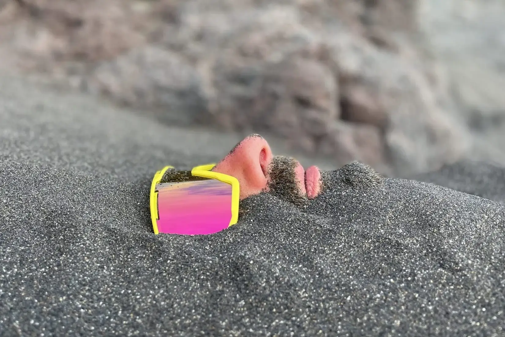living inquiry by tomm claude Ein Mann liegt im Sand eingegraben, nur seine Nase, sein Mund, sein Bart und seine reflektierende gelbe Sonnenbrille sind über der Oberfläche sichtbar - eine ruhige Szene, die an spirituelles Coaching und Zen-Meditation für emotionale Heilung und Verkörperung erinnert. Sandige Felsen bilden den Hintergrund. spirituelles coaching und zen meditation für emotionale heilung und embodiment