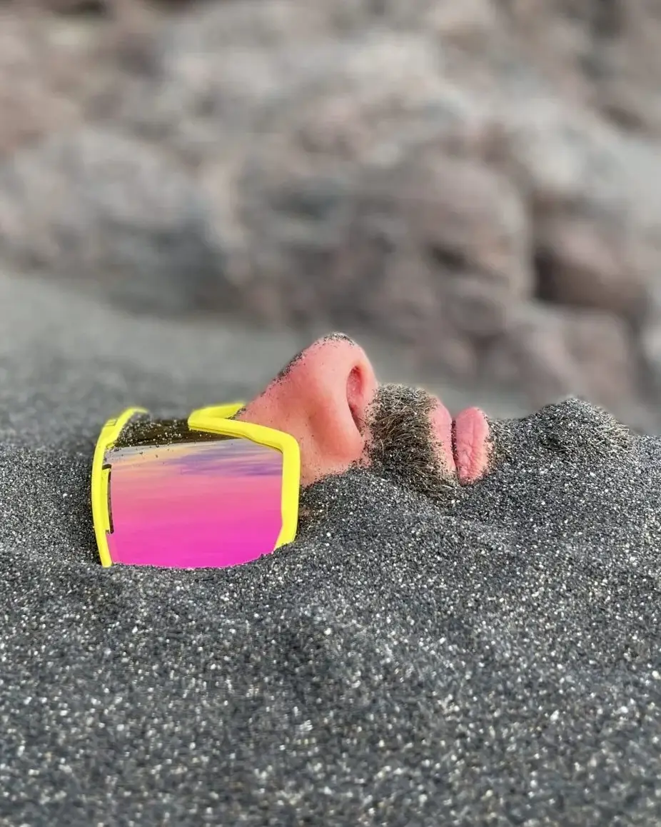 living inquiry by tomm claude Ein Mann liegt im Sand eingegraben, nur seine Nase, sein Mund, sein Bart und seine reflektierende gelbe Sonnenbrille sind über der Oberfläche sichtbar - eine ruhige Szene, die an spirituelles Coaching und Zen-Meditation für emotionale Heilung und Verkörperung erinnert. Sandige Felsen bilden den Hintergrund. spirituelles coaching und zen meditation für emotionale heilung und embodiment