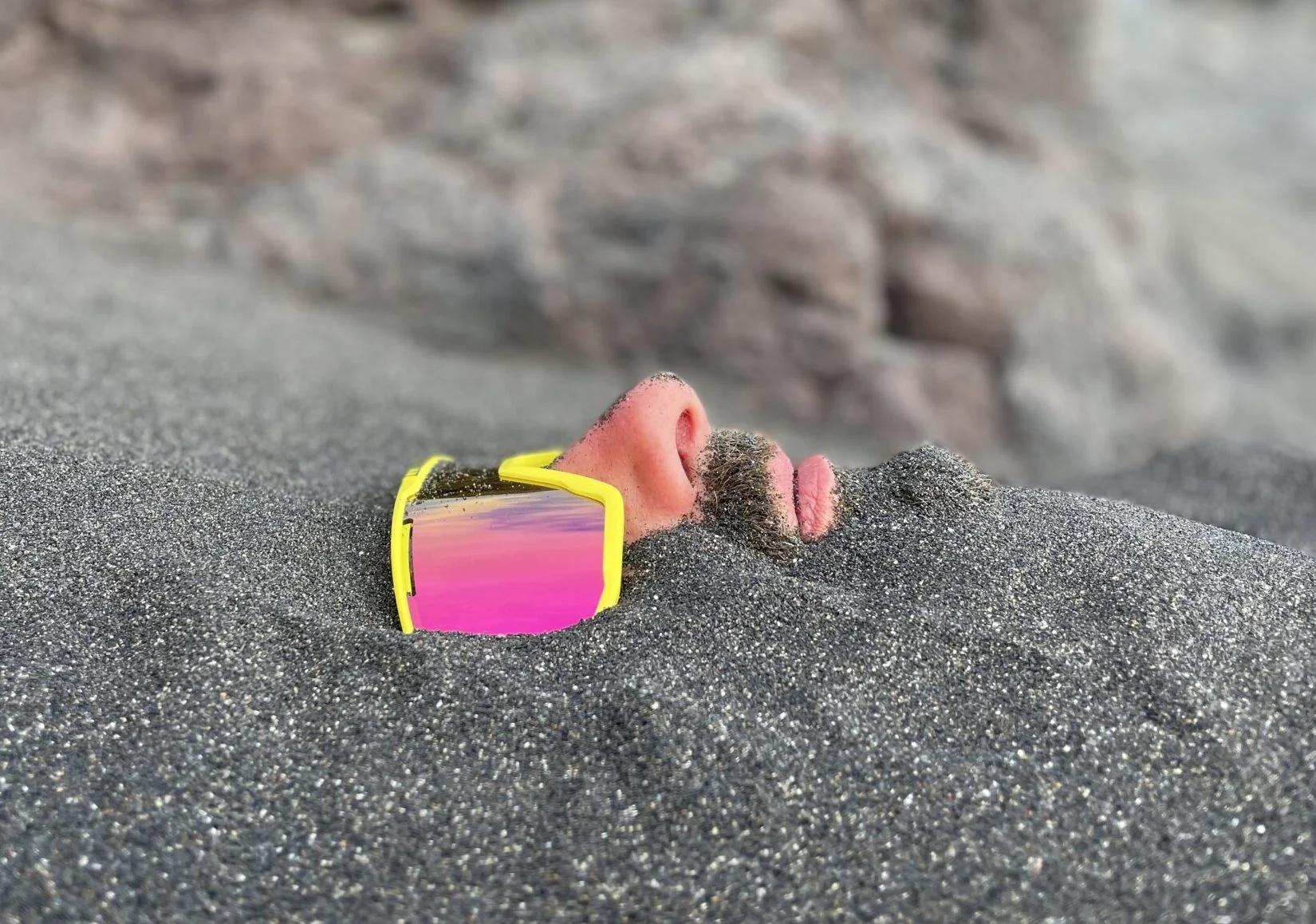living inquiry by tomm claude Ein Mann liegt im Sand eingegraben, nur seine Nase, sein Mund, sein Bart und seine reflektierende gelbe Sonnenbrille sind über der Oberfläche sichtbar - eine ruhige Szene, die an spirituelles Coaching und Zen-Meditation für emotionale Heilung und Verkörperung erinnert. Sandige Felsen bilden den Hintergrund. spirituelles coaching und zen meditation für emotionale heilung und embodiment