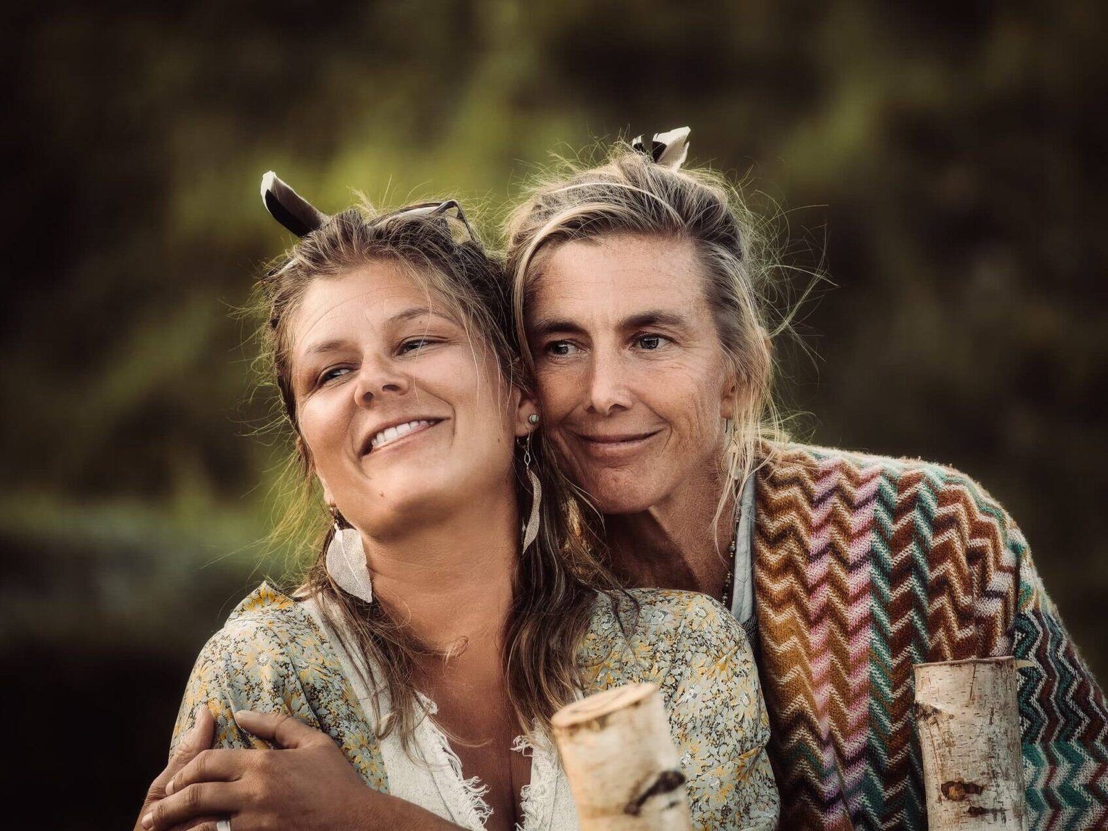living inquiry by tomm claude Zwei Frauen stehen im Freien, lächeln und umarmen sich. Beide tragen gemusterte Schals und Federohrringe. Ihre Freundschaft und Verbundenheit heben sich deutlich von dem verschwommenen grünen Laub im Hintergrund ab. spirituelles coaching und zen meditation für emotionale heilung, erwachen und essenz