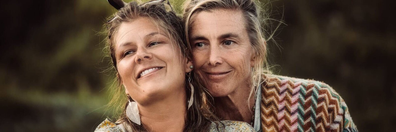 living inquiry by tomm claude Zwei Frauen stehen im Freien, lächeln und umarmen sich. Beide tragen gemusterte Schals und Federohrringe. Ihre Freundschaft und Verbundenheit heben sich deutlich von dem verschwommenen grünen Laub im Hintergrund ab. spirituelles coaching und zen meditation für emotionale heilung, erwachen und essenz