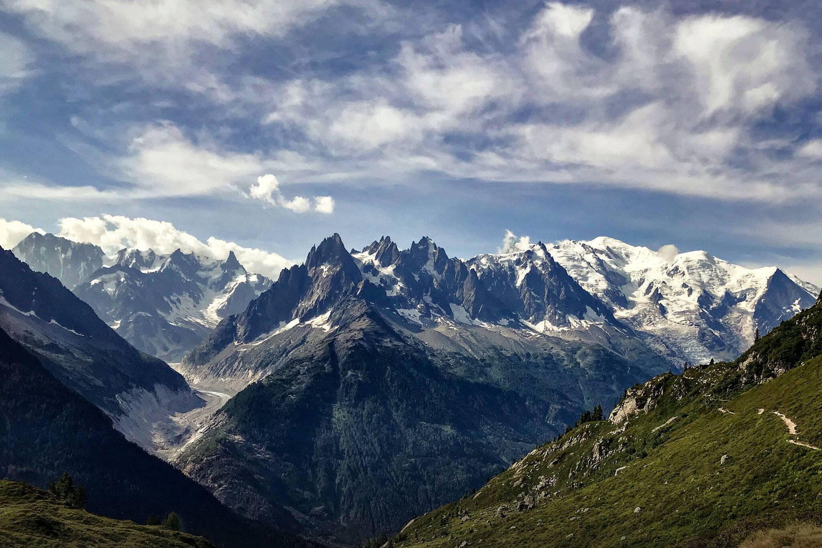 spirituelles coaching und zen meditation für emotionale heilung und embodiment Schroffe, schneebedeckte Berge erheben sich unter einem teilweise bewölkten Himmel, mit grünen Hügeln und Tälern im Vordergrund - eine Szene, die zu zenartiger Ruhe einlädt und zu spirituellem Coaching inmitten dramatischer Gipfel, die sich über den Horizont erstrecken, inspiriert.