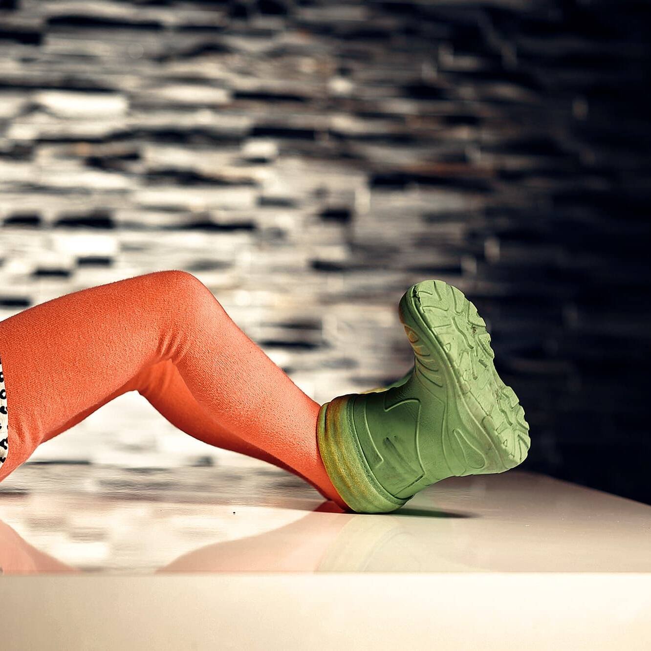 living inquiry by tomm claude Ein Kind verkörpert Freude und Einfachheit. Es liegt auf dem Rücken, trägt orangefarbene Strumpfhosen und grüne Schuhe und hat die Beine auf einer glänzend weißen Fläche angewinkelt. Der Hintergrund ist eine strukturierte, dunkle Wand; nur die untere Hälfte des Körpers ist sichtbar. spirituelles coaching und zen meditation für emotionale heilung, erwachen und essenz