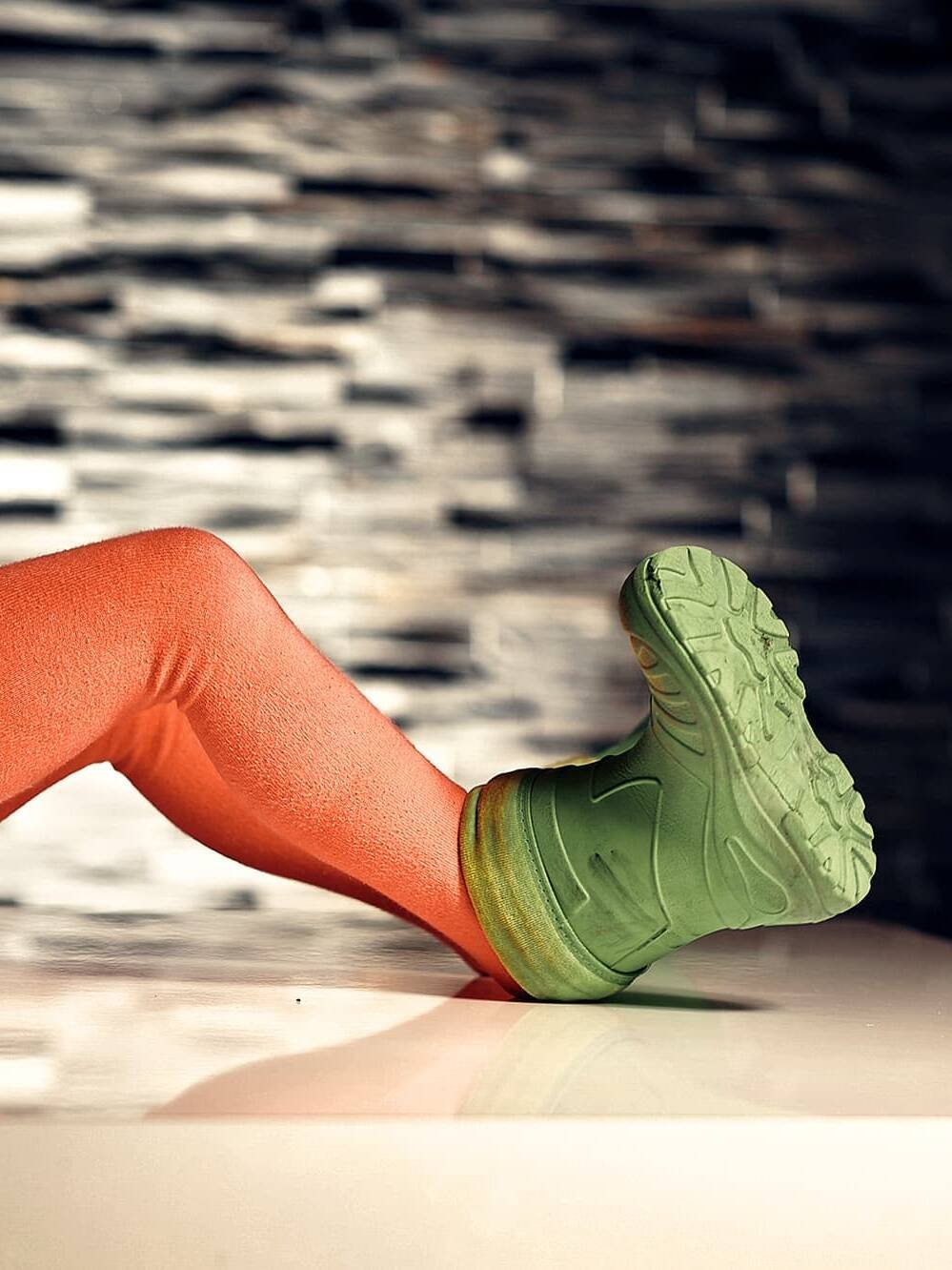 living inquiry by tomm claude Ein Kind verkörpert Freude und Einfachheit. Es liegt auf dem Rücken, trägt orangefarbene Strumpfhosen und grüne Schuhe und hat die Beine auf einer glänzend weißen Fläche angewinkelt. Der Hintergrund ist eine strukturierte, dunkle Wand; nur die untere Hälfte des Körpers ist sichtbar. spirituelles coaching und zen meditation für emotionale heilung, erwachen und essenz