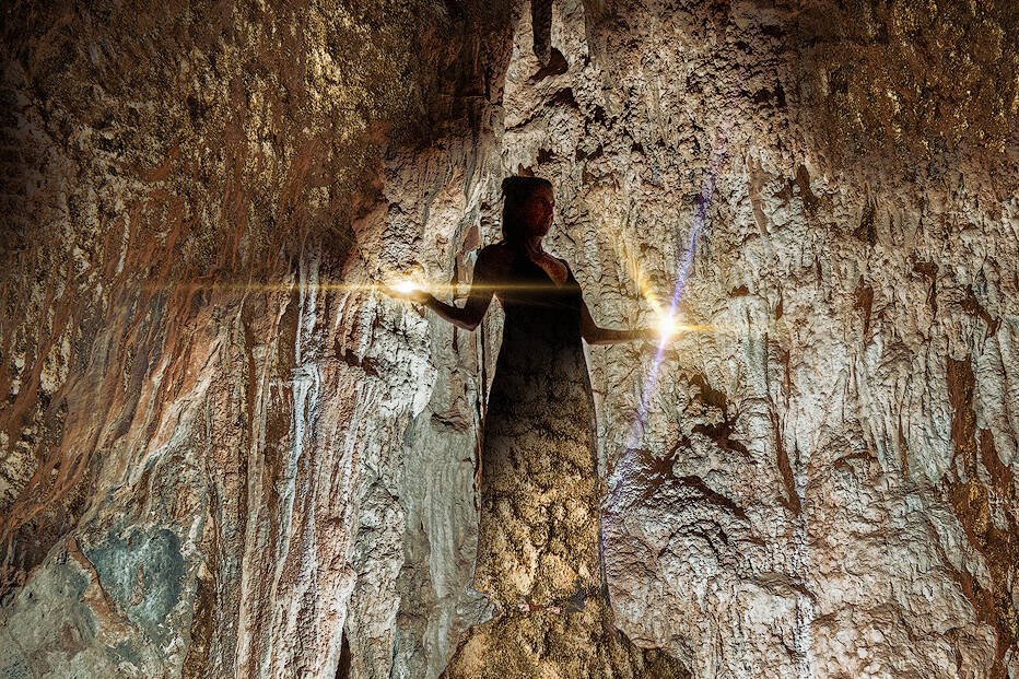 spirituelles coaching und zen meditation für emotionale heilung und embodiment Eine Person steht in einer zerklüfteten Höhle, die von einem Lichtstrahl beleuchtet wird, der eine dramatische Silhouette vor den strukturierten Felswänden erzeugt - eine Atmosphäre, die an Zazen-Meditation inmitten von zerklüfteten Kanten und erdigen Tönen erinnert.
