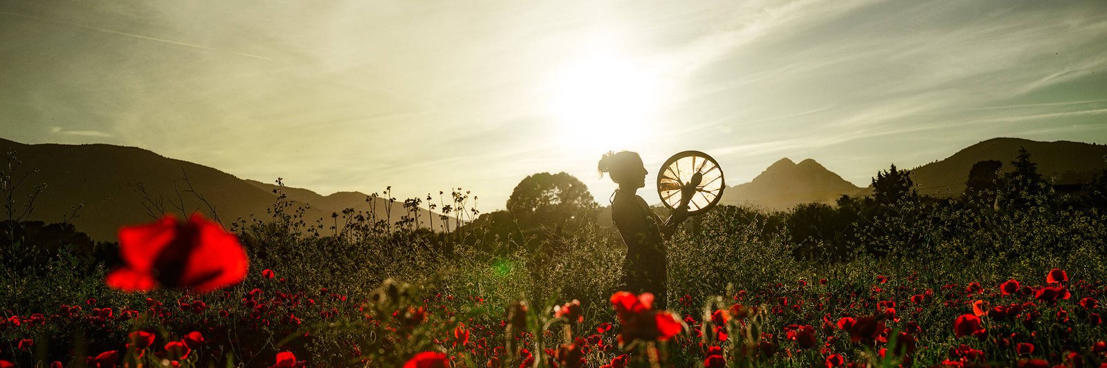 living inquiry by tomm claude Eine Frau steht bei Sonnenuntergang in einem Feld voller roter Mohnblumen und schlägt eine schamanische Trommel. Die helle Sonne schafft eine dramatische Silhouette, die an Einfachheit und Freude erinnert, mit Bergen und Bäumen im Hintergrund. spirituelles coaching und zen meditation für emotionale heilung und embodiment