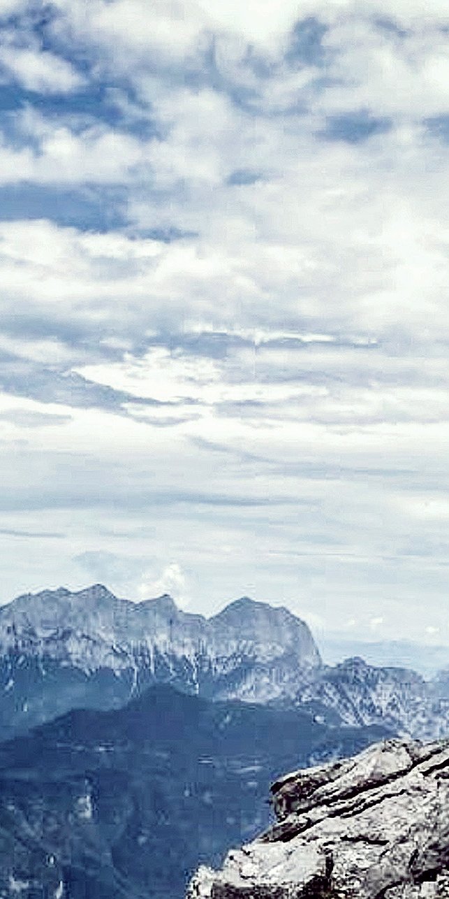 Eine Person steht auf einer Felsklippe und genießt eine innere Reise, während sie unter einem wolkigen Himmel auf ferne Berge blickt. Die Szene erinnert an Gewahrsein und verbindet Abenteuer und Einsamkeit in dieser hochgelegenen, natürlichen Umgebung.