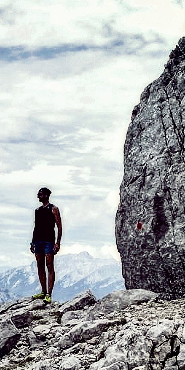 Eine Person steht auf einer Felsklippe und genießt eine innere Reise, während sie unter einem wolkigen Himmel auf ferne Berge blickt. Die Szene erinnert an Gewahrsein und verbindet Abenteuer und Einsamkeit in dieser hochgelegenen, natürlichen Umgebung.