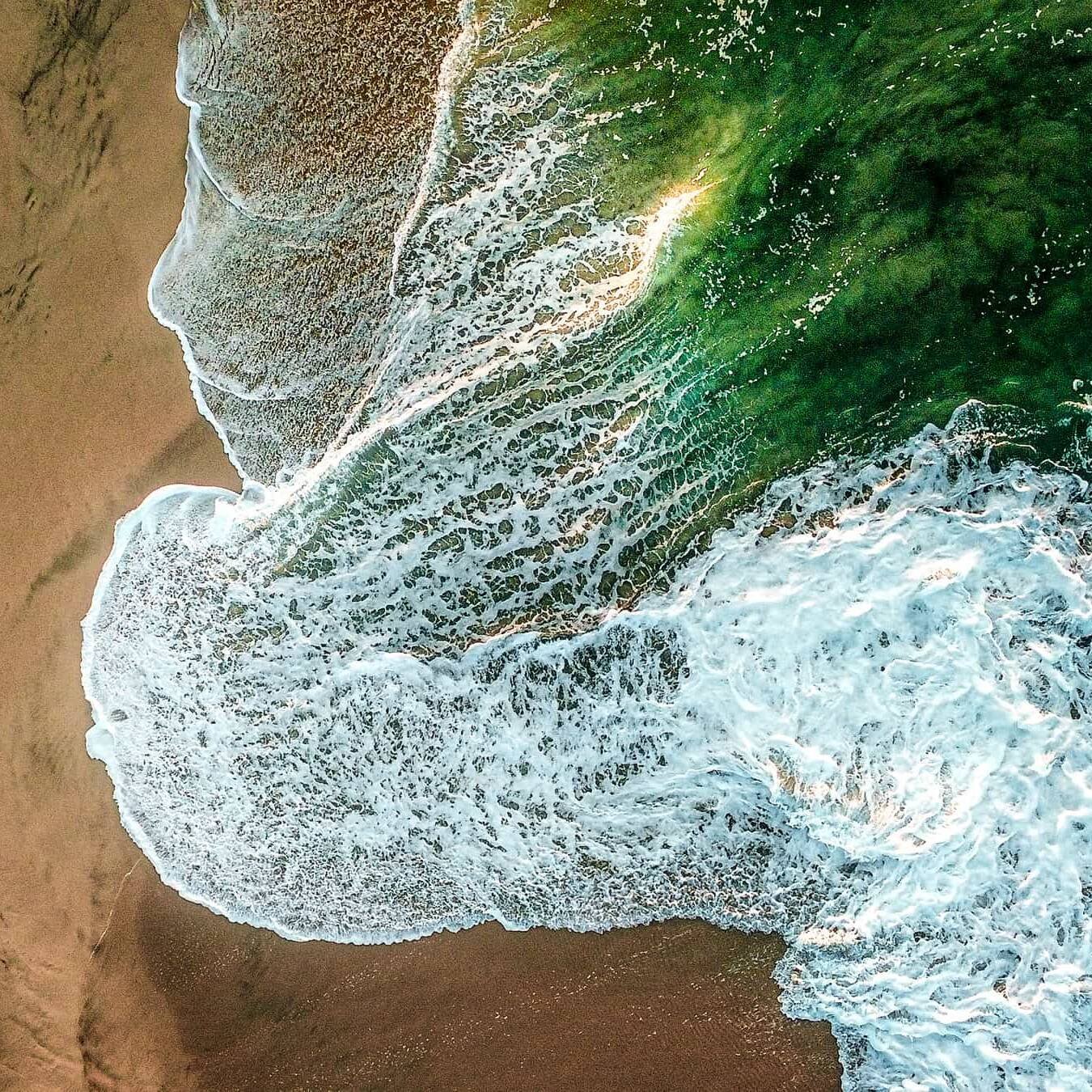 living inquiry by tomm claude Luftaufnahme von Wellen, die auf einen Sandstrand prallen und die Ruhe von spirituelles coaching und zen meditation für emotionale heilung und embodiment heraufbeschwören, mit weißer Gischt, die in das dunkelgrüne Meer und den nassen braunen Sand auf der linken Seite übergeht. spirituelles coaching und zen meditation für emotionale heilung und embodiment
