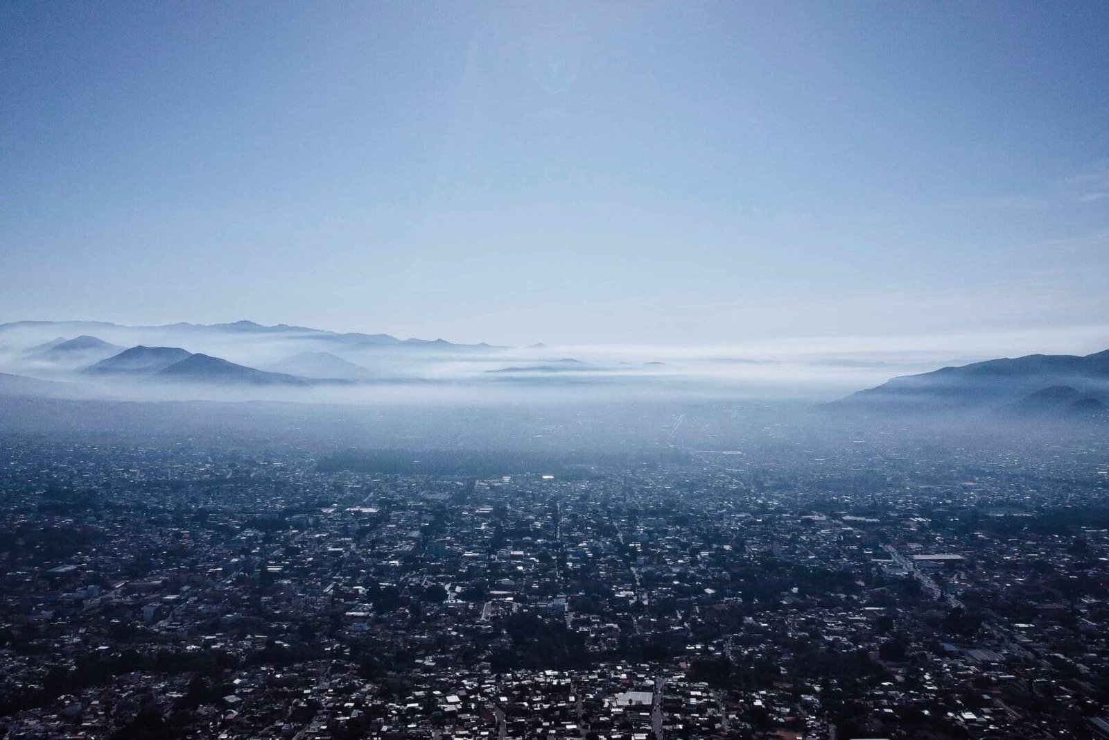 living inquiry by tomm claude Luftaufnahme einer Stadt mit einer dichten urbanen Landschaft, umgeben von Bergen unter einem dunstigen Himmel. Nebel liegt zwischen den Hügeln und spiegelt das ruhige Gleichgewicht zwischen Grenzen setzen und Selbstentdeckung in dieser ruhigen, weitläufigen Szene wider. spirituelles coaching und zen meditation für emotionale heilung, erwachen und essenz