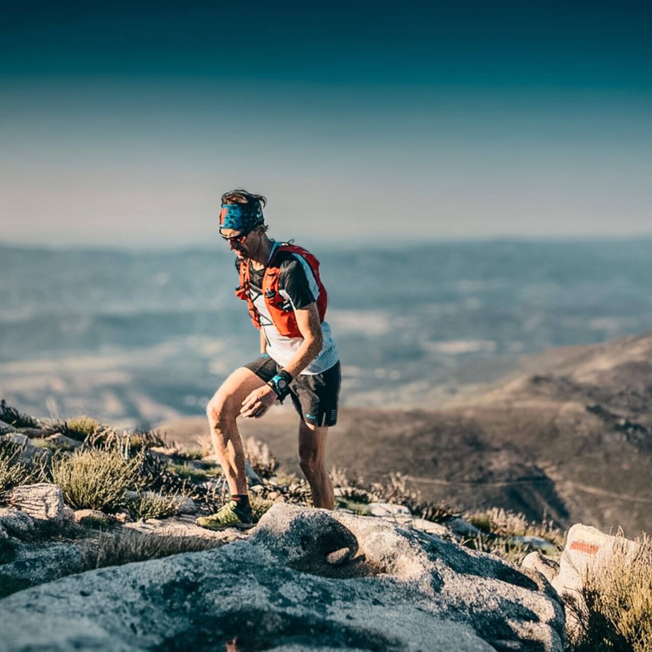 living inquiry by tomm claude Ein Trailrunner mit Outdoor-Ausrüstung, Trinkrucksack und Sonnenbrille erklimmt einen felsigen Berghang und schiebt sich durch die Selbstzweifel, während sich im Hintergrund malerische Hügel und ein klarer Himmel erstrecken. spirituelles coaching und zen meditation für emotionale heilung, erwachen und essenz