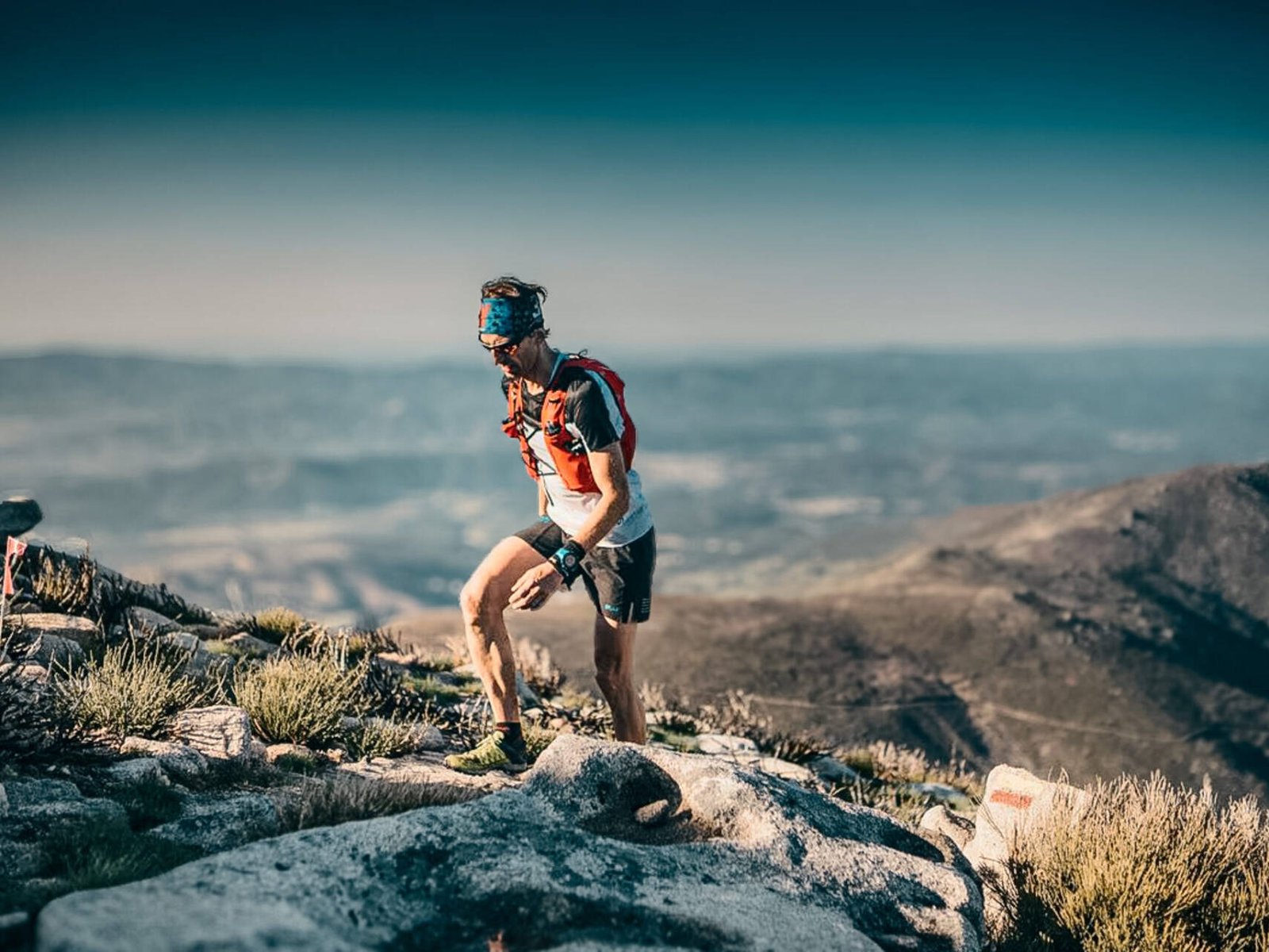 living inquiry by tomm claude Ein Trailrunner mit Outdoor-Ausrüstung, Trinkrucksack und Sonnenbrille erklimmt einen felsigen Berghang und schiebt sich durch die Selbstzweifel, während sich im Hintergrund malerische Hügel und ein klarer Himmel erstrecken. spirituelles coaching und zen meditation für emotionale heilung, erwachen und essenz