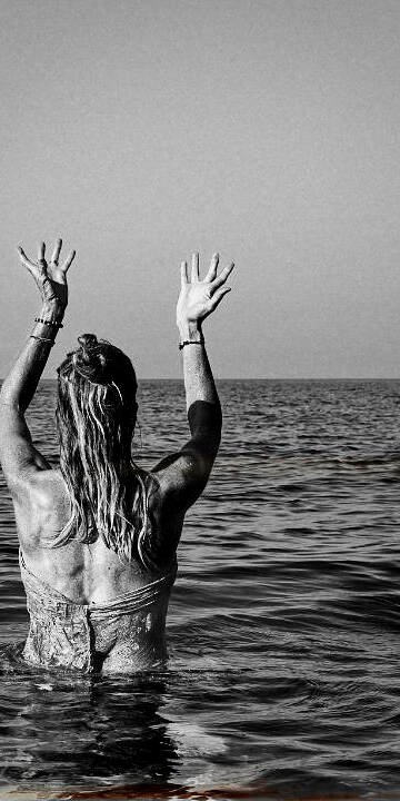 living inquiry by tomm claude Eine lächelnde Frau steht hüfttief im Meer und hält spielerisch ein Kleidungsstück hoch, während sie in die Kamera schaut. Sie verkörpert innere Ruhe und Freude daran, im Hier und Jetzt zu sein, während sich hinter ihr der Himmel und der Horizont erstrecken. spirituelles coaching und zen meditation für emotionale heilung, erwachen und essenz